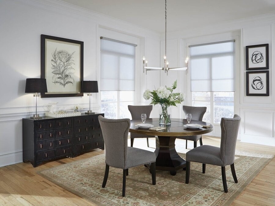 A dining room with a dining table and seats with two windows in the background featuring motorized shades, with a smart lighting fixture over the table and two on a cabinet against the wall.